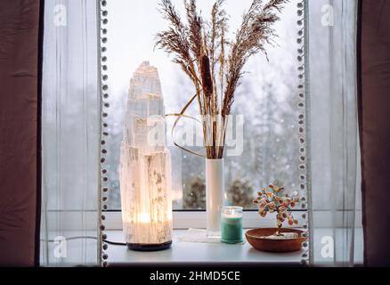 Ruvida grande selenite torre palo lampada illuminata sulla soglia della finestra di casa, spirituale decorazione accento casa. Inverno foresta sullo sfondo. Foto Stock