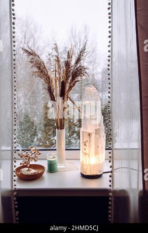 Ruvida grande selenite torre palo lampada illuminata sulla soglia della finestra di casa, spirituale decorazione accento casa. Inverno foresta sullo sfondo. Foto Stock