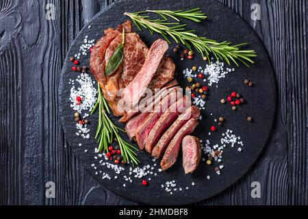 Bistecca di manzo arrosto su ardesia nera con rosmarino, sale e pepe in grani, vista dall'alto, primo piano Foto Stock