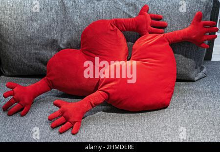 Due morbidi cuscini rossi a forma di cuore con le mani su un divano grigio. Foto Stock