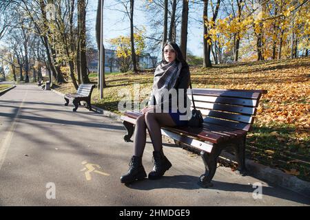 giovane ragazza brunetta in giacca nera e abito blu siede sulla panchina del parco cittadino il giorno d'autunno Foto Stock