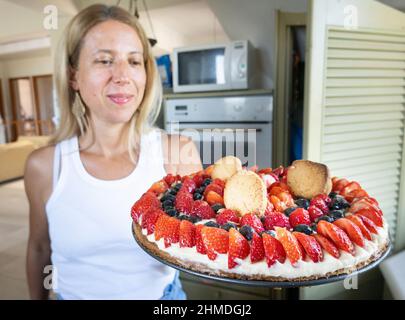 Bella donna con crostata guarnita con panna montata, bacche fresche e biscotti in cucina Foto Stock