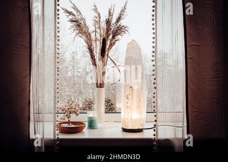 Ruvida grande selenite torre palo lampada illuminata sulla soglia della finestra di casa, spirituale decorazione accento casa. Inverno foresta sullo sfondo. Foto Stock