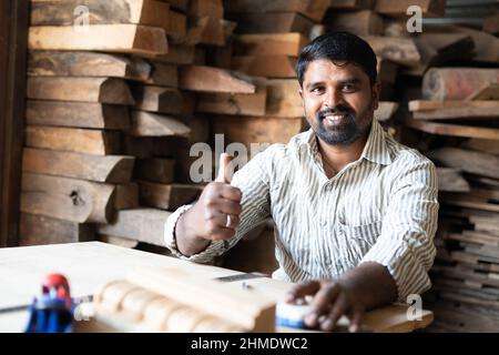 Carpentiere sorridente che mostra i pollici in su guardando la macchina fotografica mentre lavora al negozio - concetto di approvazione, promozione, artigianato e lavorazione del legno. Foto Stock