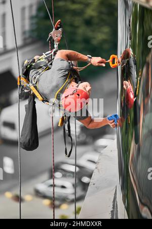 Alpinismo industriale professionale pulitore in casco protettivo appeso su corda di arrampicata di sicurezza e lavaggio facciata dell'edificio. Operatore addetto alla pulizia che utilizza un'attrezzatura di sollevamento di sicurezza. Foto Stock