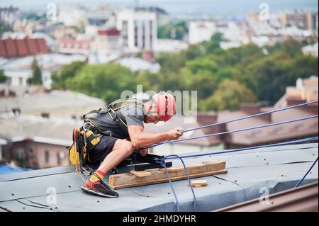 Operatore di alpinismo industriale in casco protettivo utilizzando la fune di sicurezza durante l'arrampicata sul tetto dell'edificio. Uomo che utilizza attrezzature di arrampicata di sicurezza durante lavori industriali in alto. Foto Stock