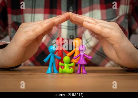 Concetto di assicurazione di famiglia con figurine di famiglia colorate coperte da mani su un tavolo di legno. Sicurezza e supporto Foto Stock