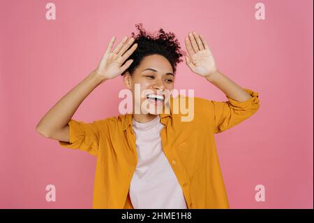 Vittoria, celebrazione, gioia. Felice bella ragazza millenaria afroamericana, ballare alla musica preferita, divertirsi, celebrare la vittoria, successo raggiunto, si leva su isolato sfondo rosa, sorridente Foto Stock