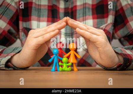 Concetto di assicurazione di famiglia con figurine di famiglia colorate coperte da mani su un tavolo di legno. Sicurezza e supporto Foto Stock