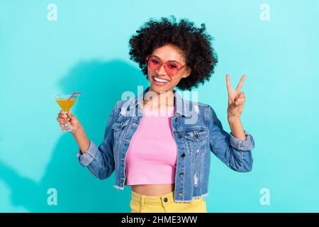 Foto di divertente bella giovane donna indossare jeans giacca occhiali v-segno bere cocktail isolato turchese sfondo colore Foto Stock