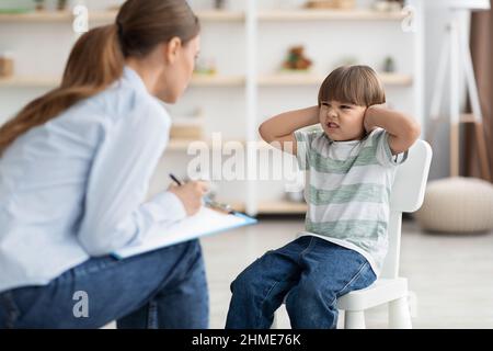 Psicoterapeuta professionista che cerca di parlare con il bambino difficile, ragazzo grumpy che copre le orecchie e il naso accigliato Foto Stock