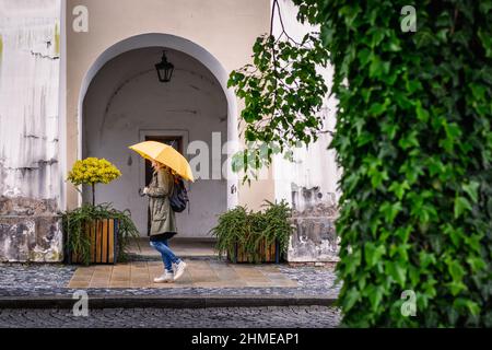 Donna con ombrello giallo che cammina per la strada della città sotto la pioggia. Passeggiate turistiche femminili nella città vecchia europea Kromeriz, Repubblica Ceca Foto Stock