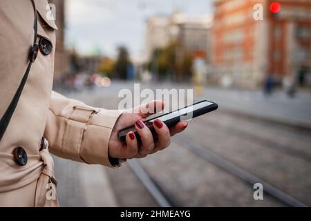Donna che controlla il suo smartphone mentre attende alla fermata dell'autobus in via cittadina. Cellulare in mano femminile Foto Stock