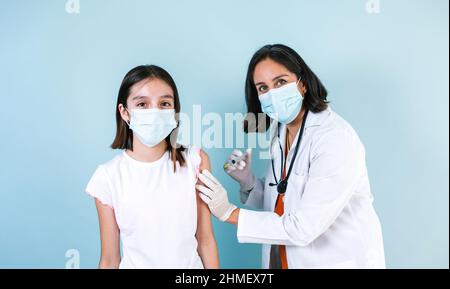 Latin medico donna medico o infermiere che dà il vaccino del coronavirus sparato a ragazza bambino ispanica su sfondo blu studio Foto Stock