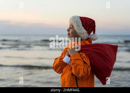 Ritratto di un ragazzo carino in un cappellino di San Nicola, con una borsa di Natale e Capodanno regali sullo sfondo del mare, il ragazzo tiene una borsa di gif Foto Stock