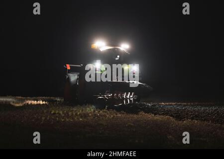 Un agricoltore che lavora a tarda sera, arando al buio utilizzando le luci del trattore Foto Stock