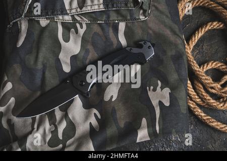 Coltello pieghevole tattico per la sopravvivenza e corda su abbigliamento militare camouflage Foto Stock