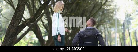 Giovane donna sorridente cammina nel parco con l'uomo in carrozzina Foto Stock