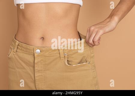 Donna con vita sottile che indossa grandi jeans color crema su sfondo di colore carne. Alimentazione sana e body building. Perdere peso mantenendo la dieta Foto Stock