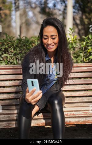 La giovane Latina sorride felicemente facendo una videochiamata utilizzando uno smartphone nel parco. Latina donna seduta su una panca utilizzando il suo smartphone. Foto Stock