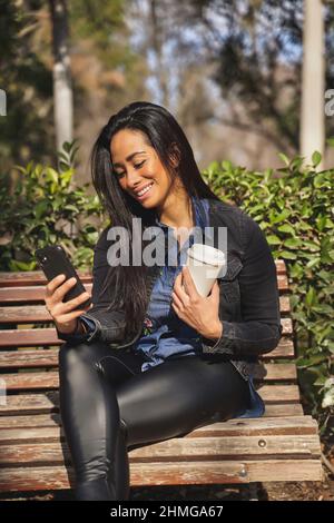 Donna latina che beve caffè e texting al parco. Foto di alta qualità Foto Stock