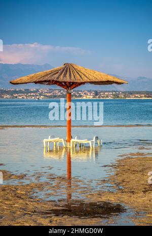 Spiaggia di sabbia di mare, ombrellone e due sdraio allagati durante la marea, nella città di Nin, Croazia, Europa. Foto Stock