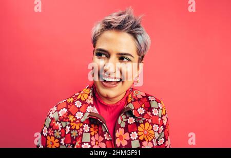 Spensierate la giovane donna che guarda via con un sorriso sul suo volto. Giovane donna felice in piedi da sola su sfondo rosso. Giovane donna alla moda con la ragazza Foto Stock