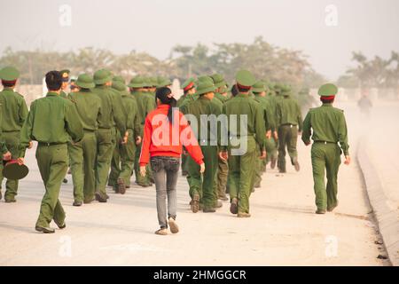 Vista posteriore della ragazza adolescente in camicia rossa che cammina con un gruppo di giovani soldati vietnamiti su una strada, accademie militari vietnamite. Vietnam. Foto Stock