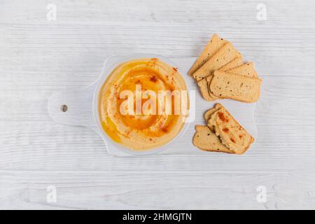 Una ciotola di ceci hummus fatto in casa con olio d'oliva, pane croccante e paprika affumicata su sfondo bianco di legno. Foto Stock