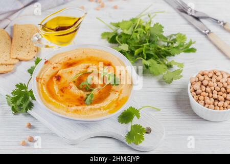 Una ciotola di ceci hummus fatto in casa con olio d'oliva, coriandolo e paprika affumicata su sfondo bianco di legno. Foto Stock