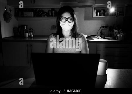 Una ragazza in una maschera e guanti impara durante la quarantena. Una donna che lavora fuori orario da casa su un notebook. Un freelance è seduto in cucina nel Foto Stock