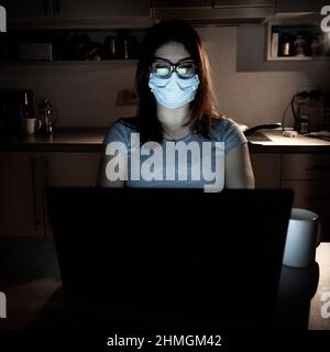 Una ragazza in una maschera e guanti impara durante la quarantena. Una donna che lavora fuori orario da casa su un notebook. Un freelance è seduto in cucina nel Foto Stock