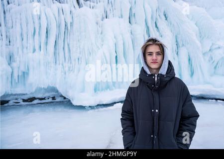 Adolescente sullo sfondo di una parete di ghiaccio, sul lago Baikal, guarda nella macchina fotografica in abiti invernali con un cappuccio. Foto Stock