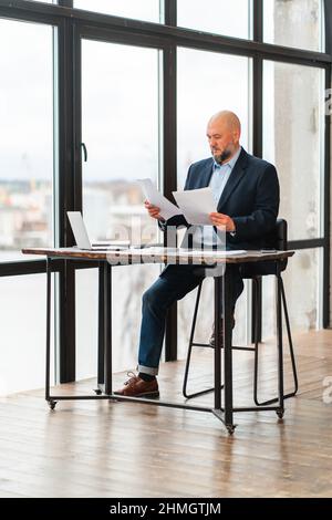 Uomo d'affari maturo alla scrivania d'ufficio che analizza i documenti e fa il lavoro di ufficio Foto Stock