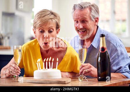 Coppia in pensione festeggia il compleanno soffiando candele con bicchiere di Champagne e torta a casa Foto Stock