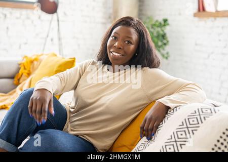 Affascinante donna afroamericana riposo a casa, serena etnia femminile seduta sul comodo divano in soggiorno moderno, sorridente e guardare la macchina fotografica. Donna calma e spensierata in abbigliamento casual Foto Stock