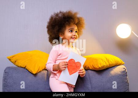 multiculturale simpatica ragazza curly tenuta cartolina con cuore disegnato in accogliente soggiorno a sera Foto Stock