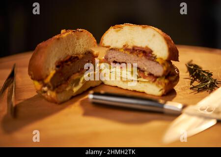 Vista ravvicinata dell'hamburger di manzo alla griglia tagliato a metà con coltelli sul tagliere. Succosa cheeseburger fatto in casa con insalata, cipolla e bastone di rosmarino su tavola di legno. Concetto di fast food. Foto Stock