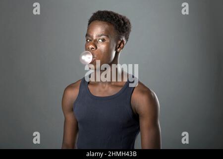 Giovane ragazzo nero in cima senza maniche che si posa su uno sfondo grigio dello studio soffiando bolle con gomma da masticare come lui sguardo lateralmente alla fotocamera Foto Stock