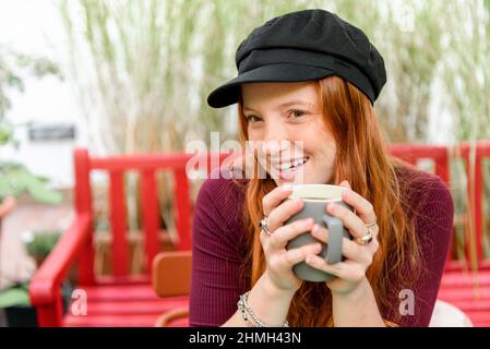 Felice giovane donna in berretto guardando via mentre si siede con una tazza di caffè su panca in giardino con piante verdi Foto Stock