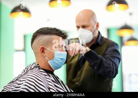 Barbiere professionale che indossa una maschera durante il Covid-19 pandemic tagliando i capelli di un giovane uomo anche in una maschera chirurgica usa e getta come da current heal Foto Stock