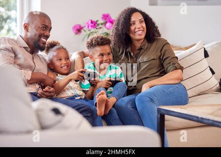I genitori seduti sul divano con i bambini a casa giocando il videogioco insieme Foto Stock
