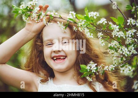 Ragazza senza denti ridente in abito bianco vicino a ciliegi in fiore. Sorriso felice e sano. Foto Stock