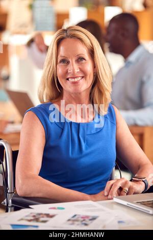 Ritratto di donna in attività matura in sedia a rotelle seduta alla scrivania che lavora sul laptop in ufficio occupato Foto Stock