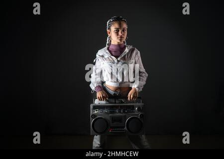 Vista frontale di una giovane donna con trecce bianche in stile urbano in piedi e tenendo in mano il registratore a nastro retrò mentre si guarda la fotocamera in studio Foto Stock