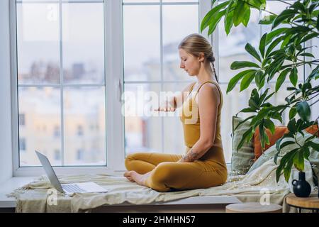 Giovane donna bionda in cima gialla e leggings medita in posa yoga seduta sul davanzale coperto di coperta di coperta e cuscini utilizzando un computer portatile Foto Stock