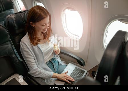 Donna sorridente che usa il computer portatile e beve champagne in aereo Foto Stock