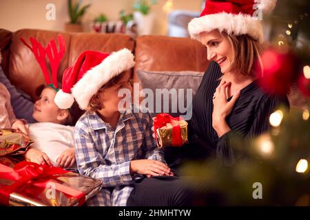 Il figlio che dà il regalo della madre come famiglia seduta sul divano a casa che apre i regali di Natale Foto Stock