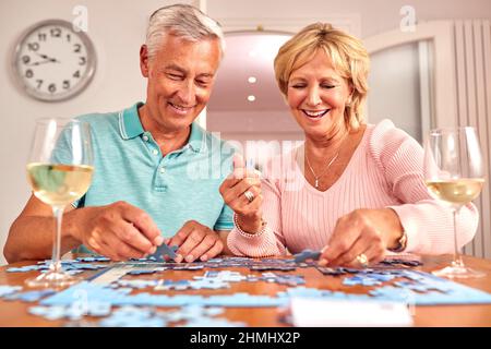 Senior coppia in pensione seduta a tavola a casa con bicchiere di vino facendo Jigsaw Puzzle Foto Stock