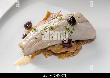 Filetto di merluzzo bollito e rosolato con carciofi e olive, primo piano su piatto bianco Foto Stock